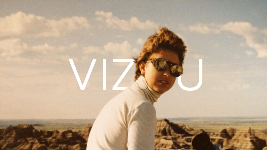 A woman in white turtleneck wearing glacier glasses sits in the foreground of the Badlands with the word Vizou behind her head